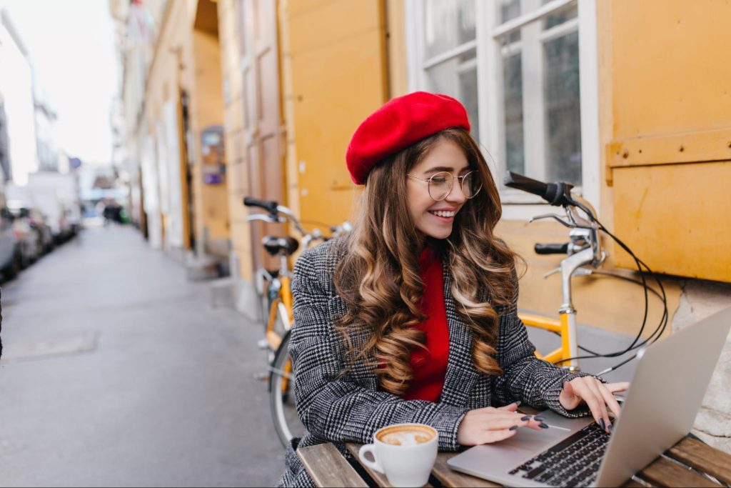 A female blogger wearing a houndstooth printed jacket and a red cap is smiling while typing on her laptop.