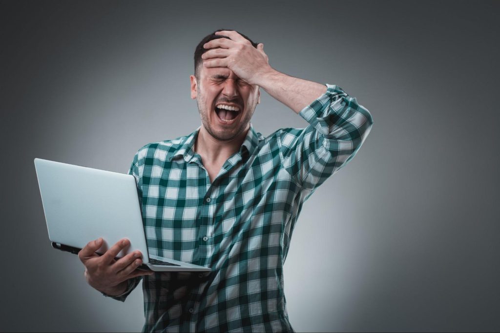 A frustrated man in a green plaid shirt holds his head in his hand while looking at a laptop with an expression of distress or overwhelm. 